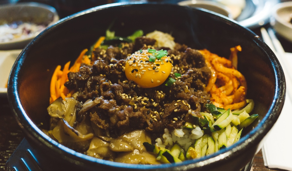 A closeup photo of a signature Korean dish called Bibimbap, consists of  beef, raw egg, namul (sautéed and seasoned vegetables) and gochujang (chili pepper paste), soy sauce, or doenjang, a fermented soybean paste.