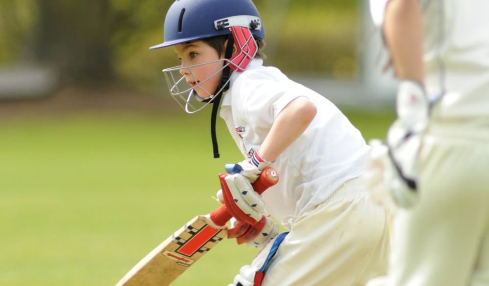 Young boy wearing protective clothing batting in a cricket match in England watched by the wicket keeper