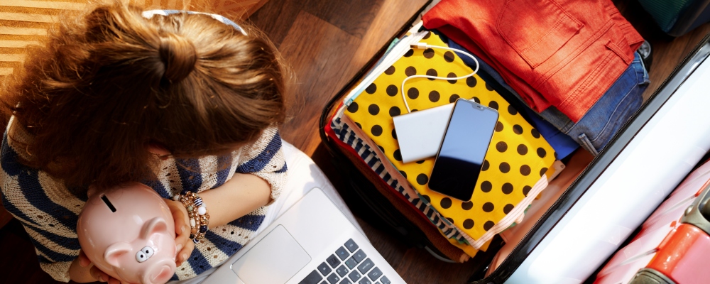 Upper view of young woman in white pants and striped blouse in the modern living room in sunny summer day holding piggy bank near open travel suitcase planning budget summer trip.