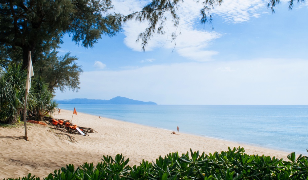 Trop[ical Phuket vibrant turqoise blue sea at Mai Khao beach with tourists, tree bush and pine tree