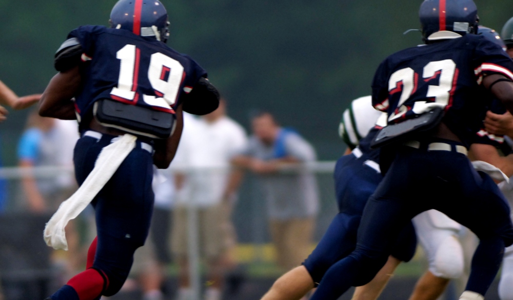 this photo is of an American Football Players Running with Football during Football Game on a football field. the football players are young men or tenagers or men playing a sport at a sporting event. they are wearing their football uniforms with football helmets during a live football game. the lighting is natural outdoor lights. and they are playing the football game on green grass or artificial turf.