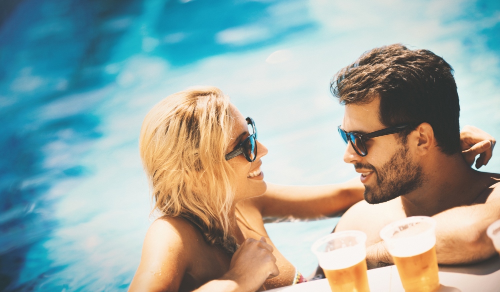 Group of young adults relaxing in a swimming pool on hot and sunny summer day. There is a guy and a girl, aged mid 20's. Both wearing sunglasses and leaning at the side of the pool. Drinking beer out of plastic cups. Side view, tilt shot.