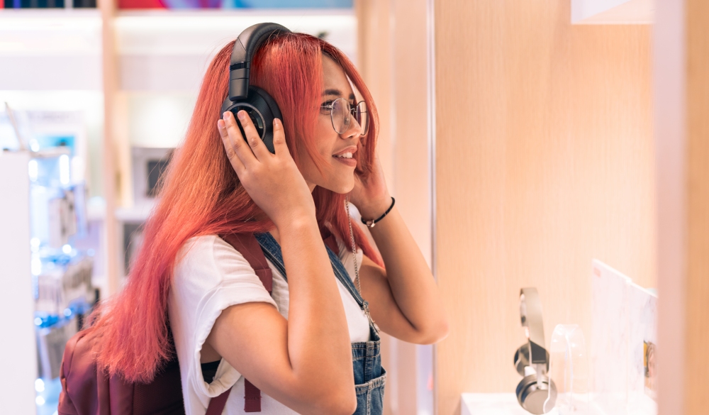 Asian social influencer woman trying on headphones inside retail store - Happy millennial diverse girl shopping & testing lifestyle music tech - Technology, electronic concept - Focus on right eye; Shutterstock ID 1443256043