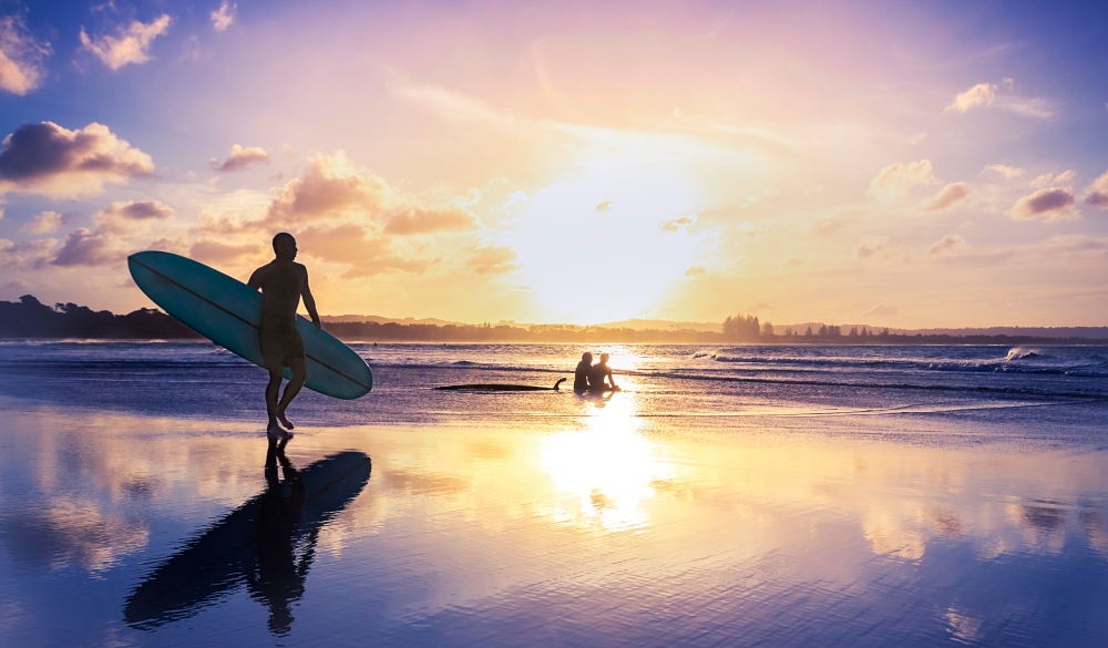 Man Walking With Surfboard By Silhouette People Sitting At Beach