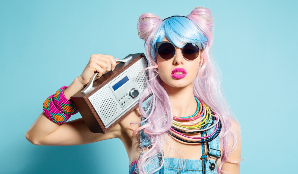 Portrait of sensual manga style blue-pink hair young woman wearing denim coveralls and sunglasses. Standing against blue background, holding radio on her shoulder. Studio shot, one person.