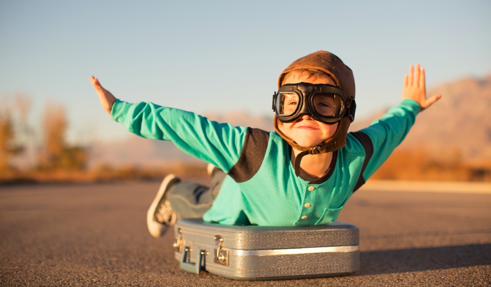 A young boy with outstretched arms lies on top of a suitcase imagining he is flying on an airplane away and traveling to exotic locations. He is wearing a flight cap and goggles and has a large smile on his face. He loves to travel.