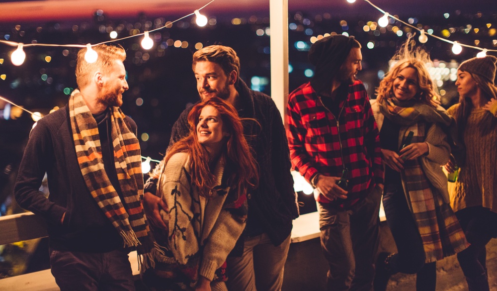 Couple of friends having a party on the rooftop of the building at sunset. Drinking bottles of beer and taking. Wearing knitted sweaters, hats and scarfs. They are happy and joyful.