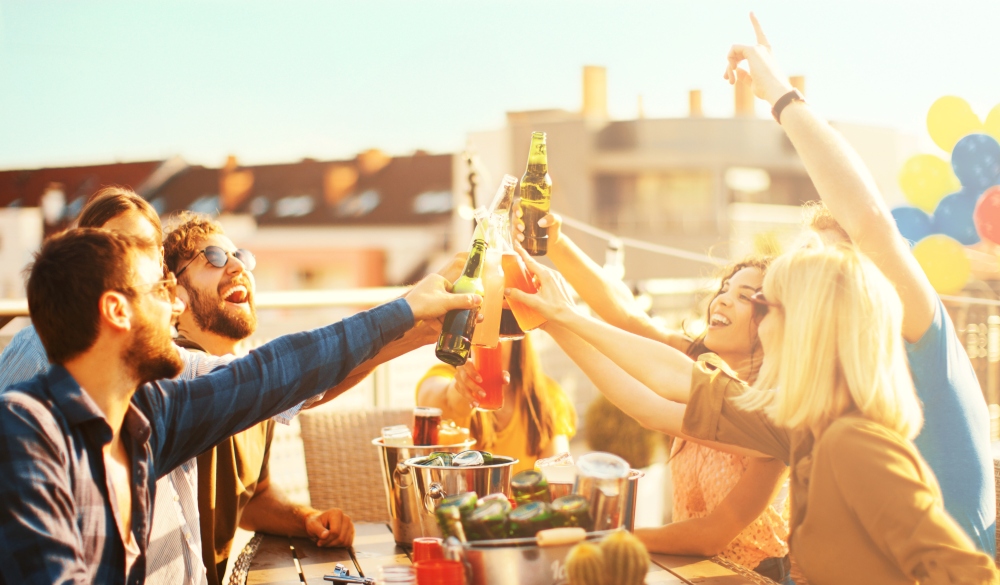 Side view of friends enjoying rooftop party. They are sitting and toasting with alcohol drinks during beautiful summer afternoon.