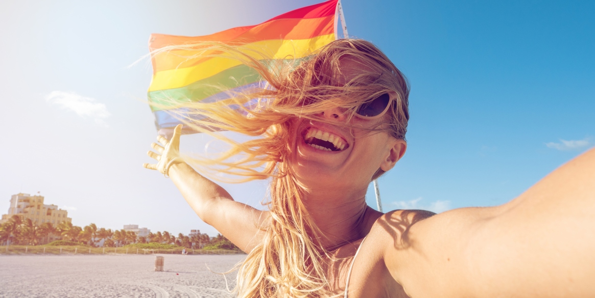 irl on beach taking selfie photo standing near rainbow gay flag on beach