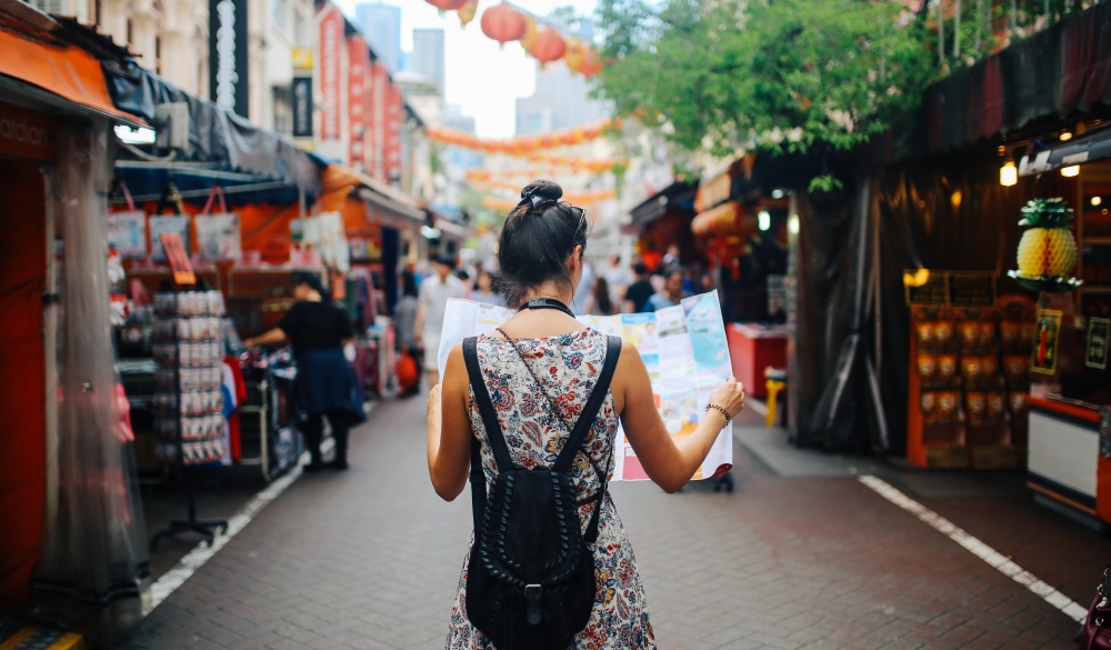 Rear view image of a young brunette woman. She is enjoying the walk and exploring the city, wearing a casual but fashionable dress, sightseeing and shopping on the Singapore street market. She is holding a large city map, checking out where to go next.