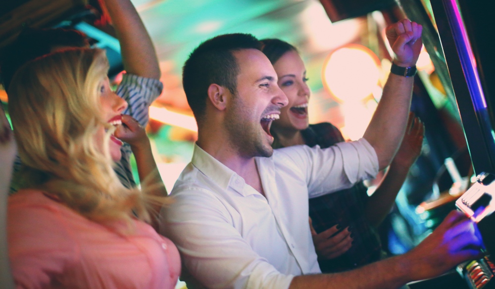 Group of young adults in mid 20's playing slots in a casino and having fun.Cheering,laughing...two couples on double date.Many slot machines stretch blurry in background.