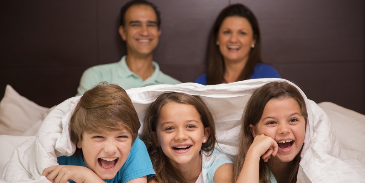 family on vacation in a hotel room or at home in bedroom. The children enjoy piling up together under the bed covers