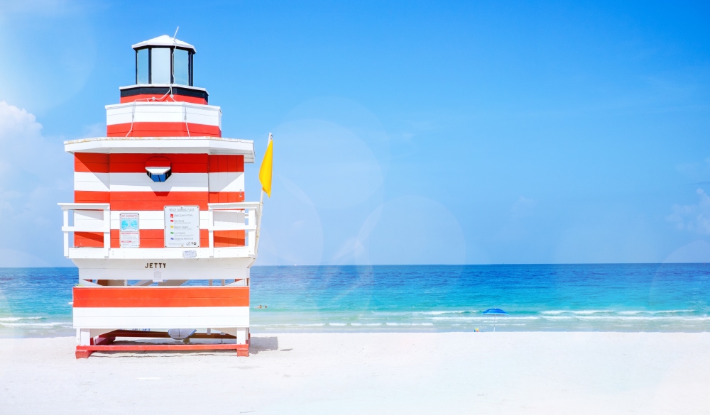 A red and white lifeguard hut, life guard station (designed like a lighthouse) with a yellow flag on an empty beach on a sunny day with clear blue sky.