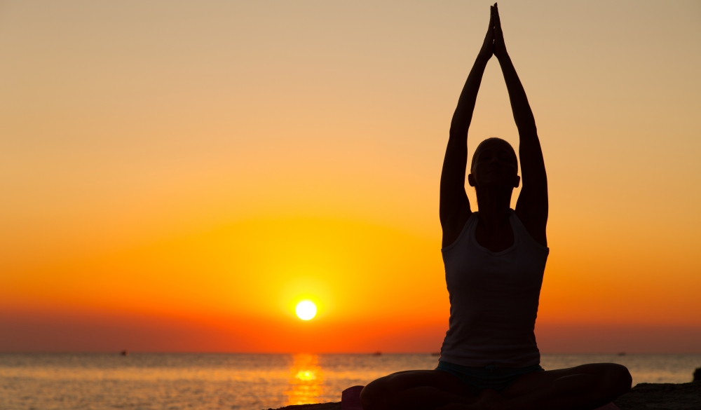 Silhouette of a young woman meditating by the sea at the sunset. Rear view. Copy space.