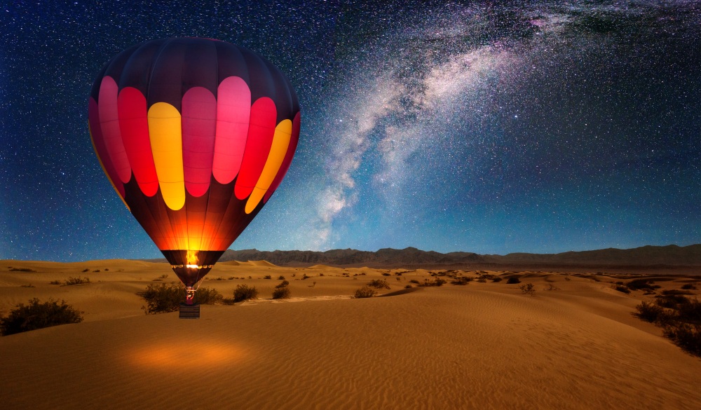 A majestic hot air balloon soars under the stars of the Milky Way, over the desert - Mesquite Dunes of Death Valley National Park. Moonlight provides luminosity showing the patterns and shapes of the desert landscape.
