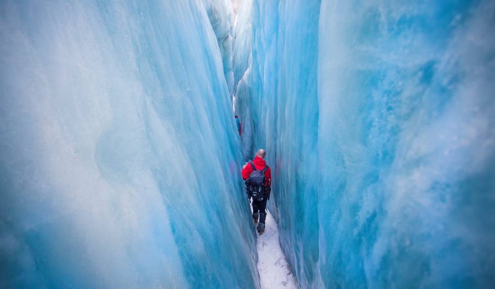 Travelers explore New Zealand's famous Franz Josef Glacier. Blue Ice, deep crevasses, caves and tunnels mark the ever changing ice. A man walks through a giant Ice crevasse, south island road trip destination