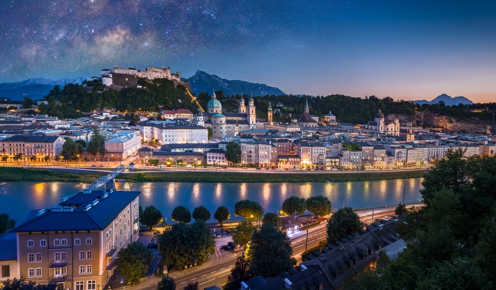 View of cityscape of Salzburg Cathedral, Fortress Hohensalzburg, and old castle in center of old town with river and road along the river at sunset time with milky way in Salzburg, Austria, Europe and also view of snow on alps mountain in background, destination for road trips in austria