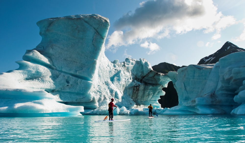 Two adults on stand up paddle board (SUP) observe hole melted in iceberg on Bear Lake in Kenai Fjords National Park, Alaska.