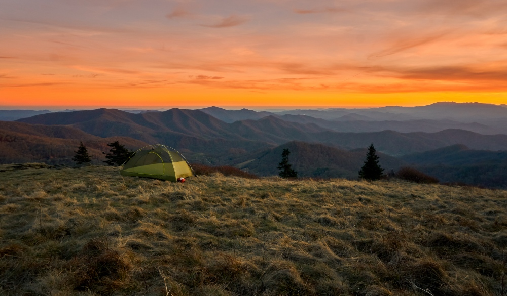 Vibrantly colorful vista of a small green tent camps on a grassy bold meadow of the Appalachian Trail in the golden sunset with blue misty mountains in the background., best hikes in the US