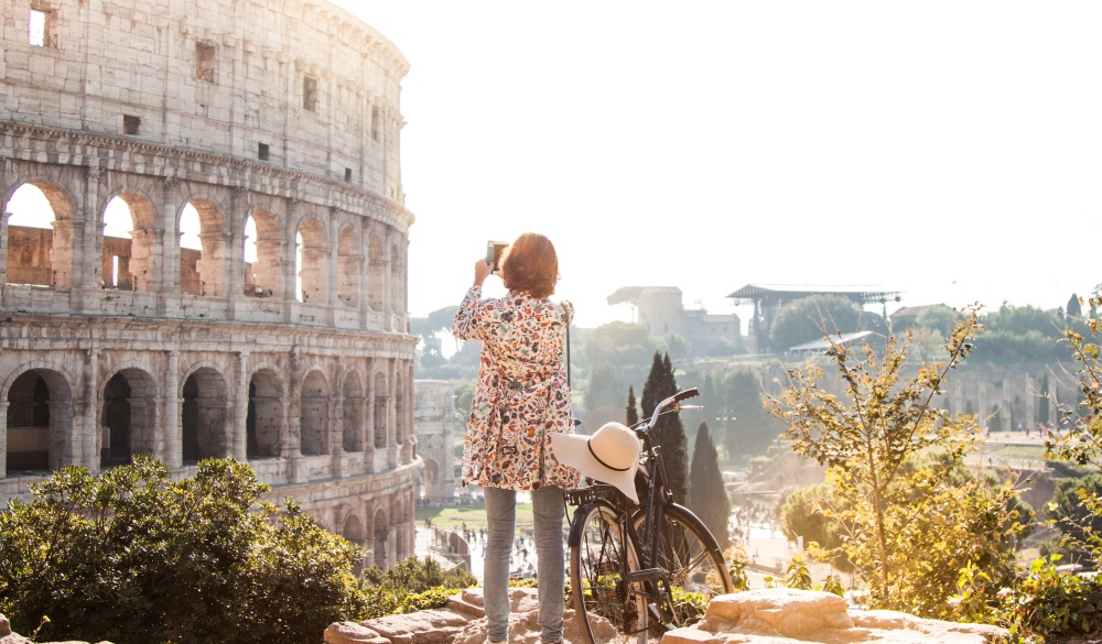 Beautiful young woman in colorful fashion dress alone on a hill with bike takes pictures of colosseum in Rome using smartphone camera at sunset. Attractive tourist girl with elegant straw hat