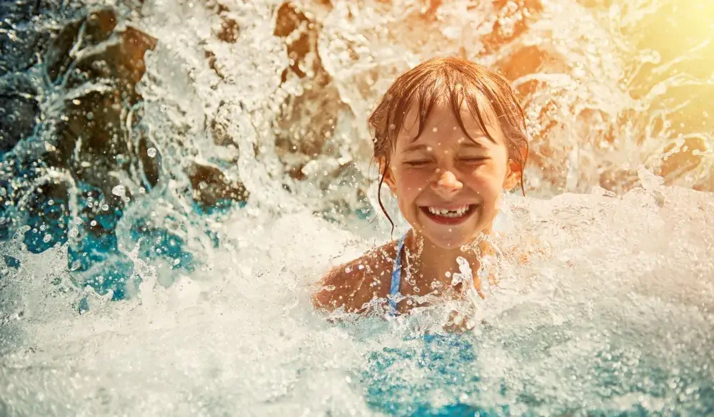 Little girl in waterpark pool being splashed by waterfall. Sunny summer day.