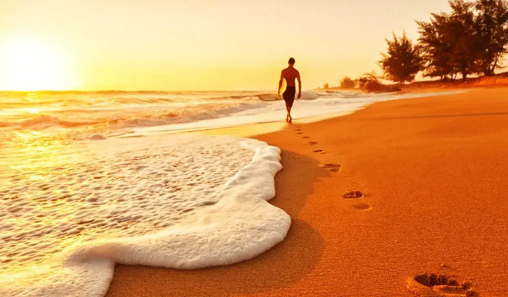Photo of footprints in a sandy Hawaiian beach at sunset, leading to the silhouette of a surfer walking into the water with his surf board; Kauai, HI.
