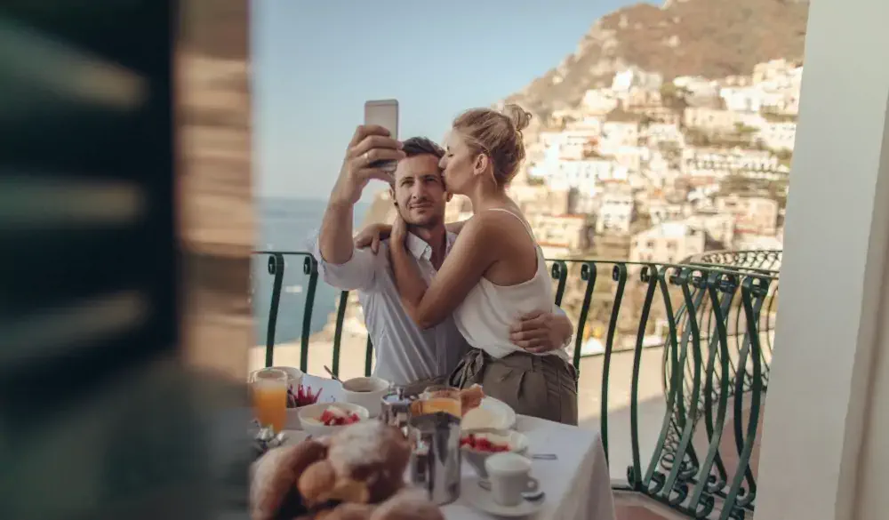 Photo of a beautiful loving couple having breakfast on the terrace with a view of amazing Positano, Italy, and taking a selfie to capture their magic moment