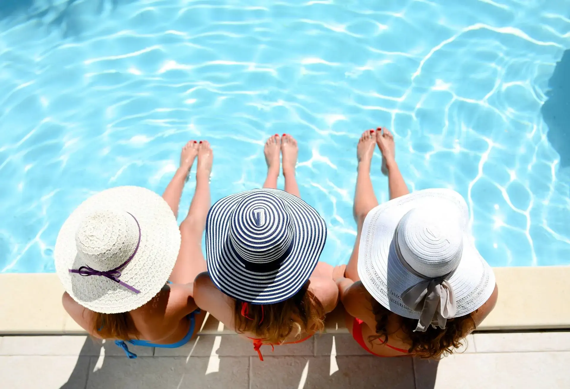 Three beautiful young woman with swimsuit and sun hat sitting by the poolside of resort swimming pool during summer holiday.