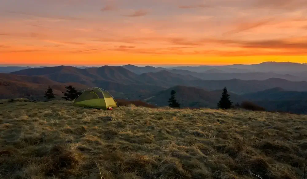 Vibrantly colorful vista of a small green tent camps on a grassy bold meadow of the Appalachian Trail in the golden sunset with blue misty mountains in the background., best hikes in the US
